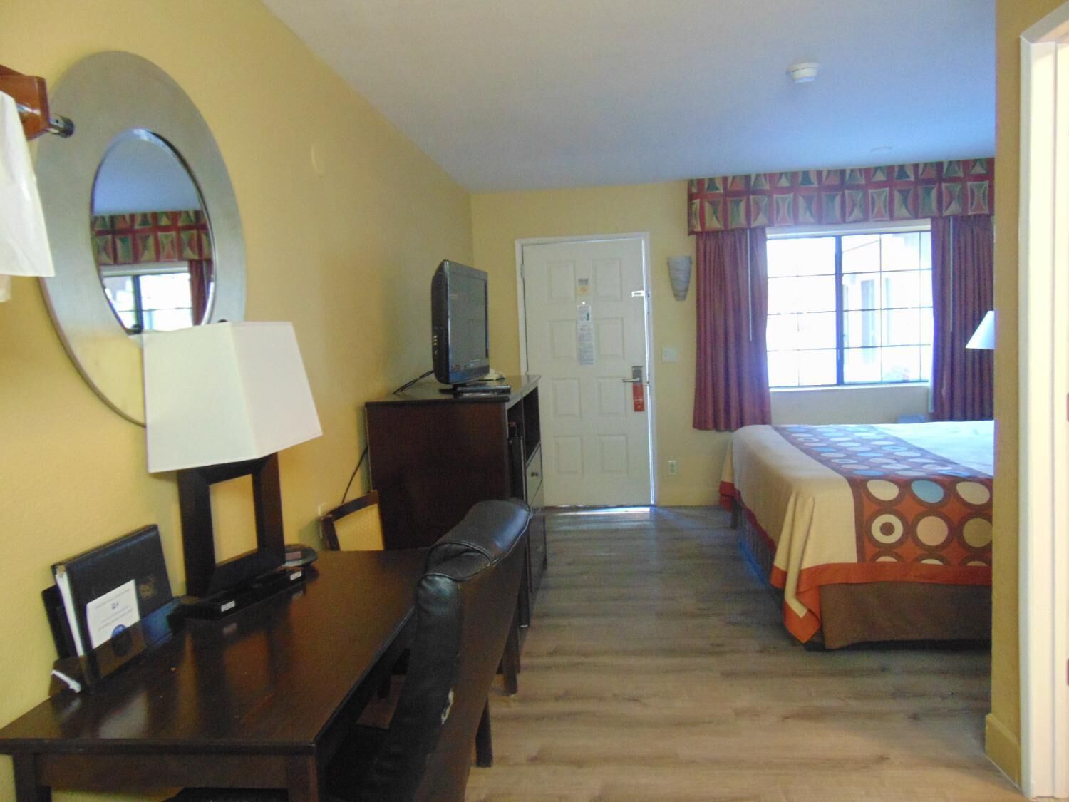 A hotel room with a bed, a desk with a chair, a television on a dresser, a mirror, and a window with curtains.