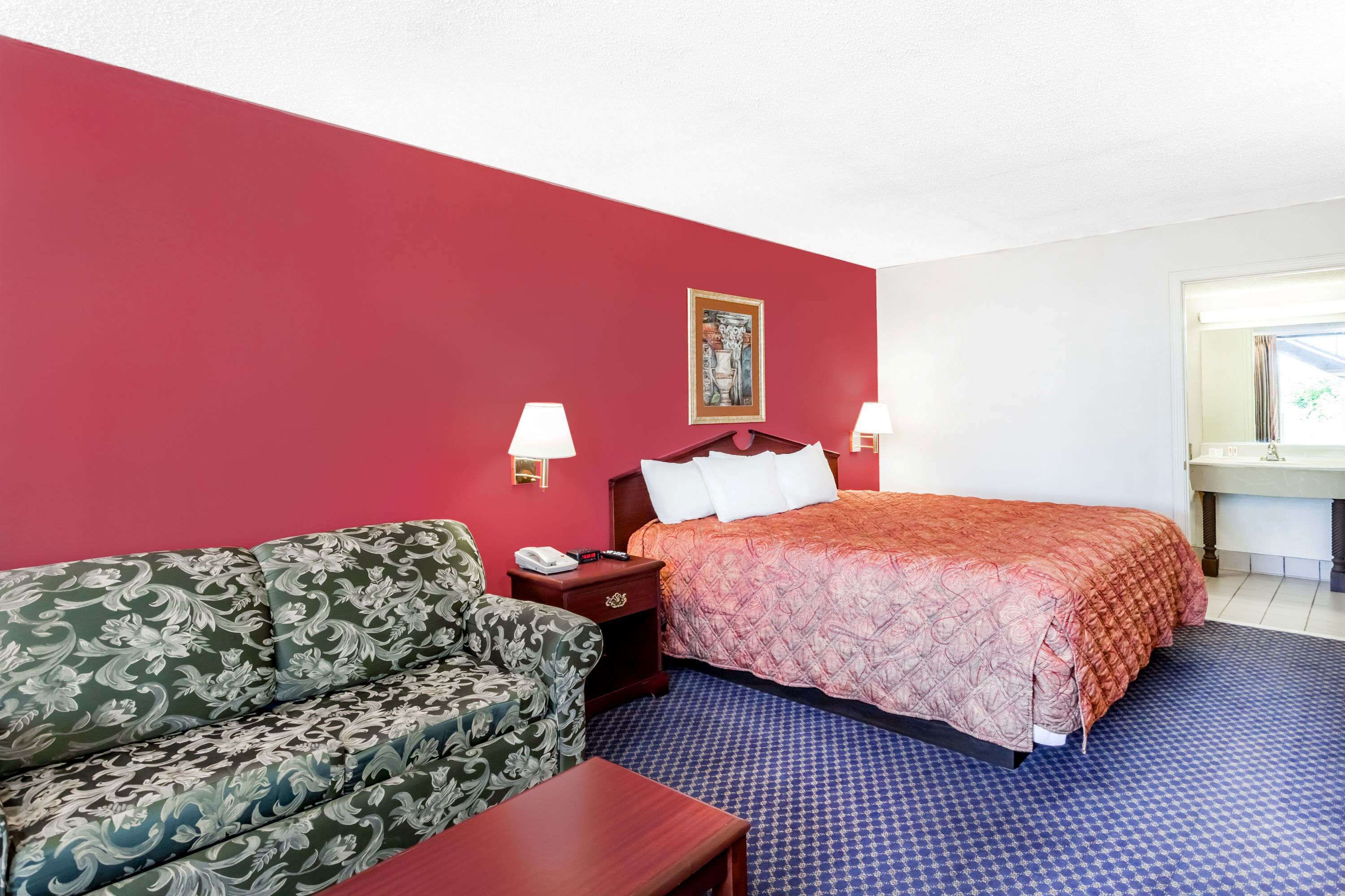 A hotel room with a red wall, a bed with a patterned bedspread, a green floral sofa, a nightstand with a lamp, and a window with a view.