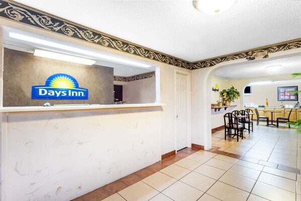 A Days Inn reception area with a marble counter, a 'Welcome' sign, and a view of the dining area with chairs and a table.