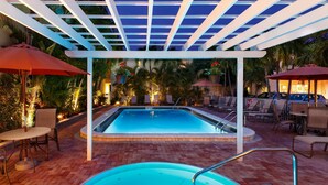 Una piscina con una pérgola blanca sobre ella, rodeada de sillones de descanso y sombrillas.