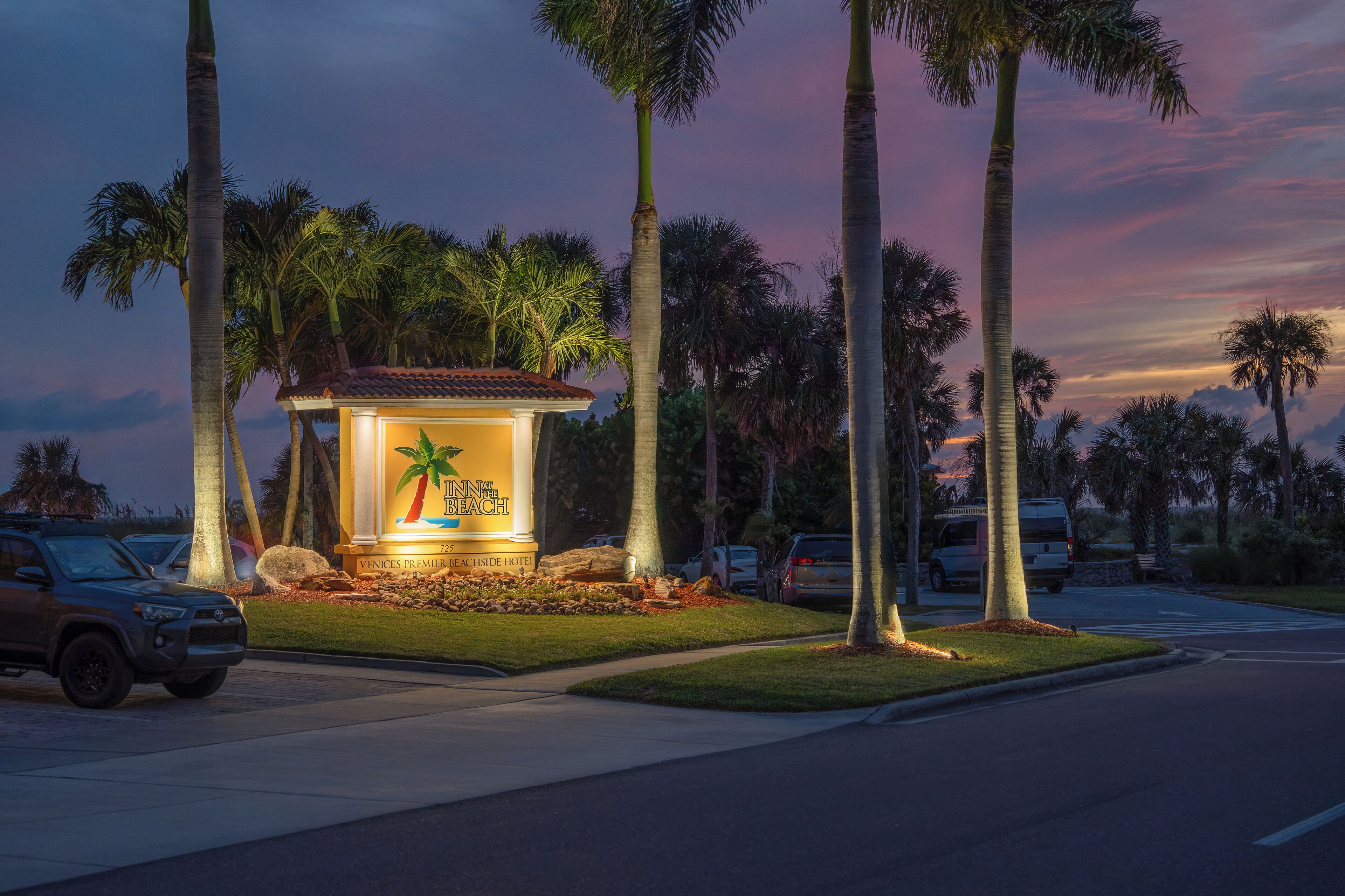A well-lit hotel sign with palm trees and a beach theme, surrounded by parked cars and palm trees.