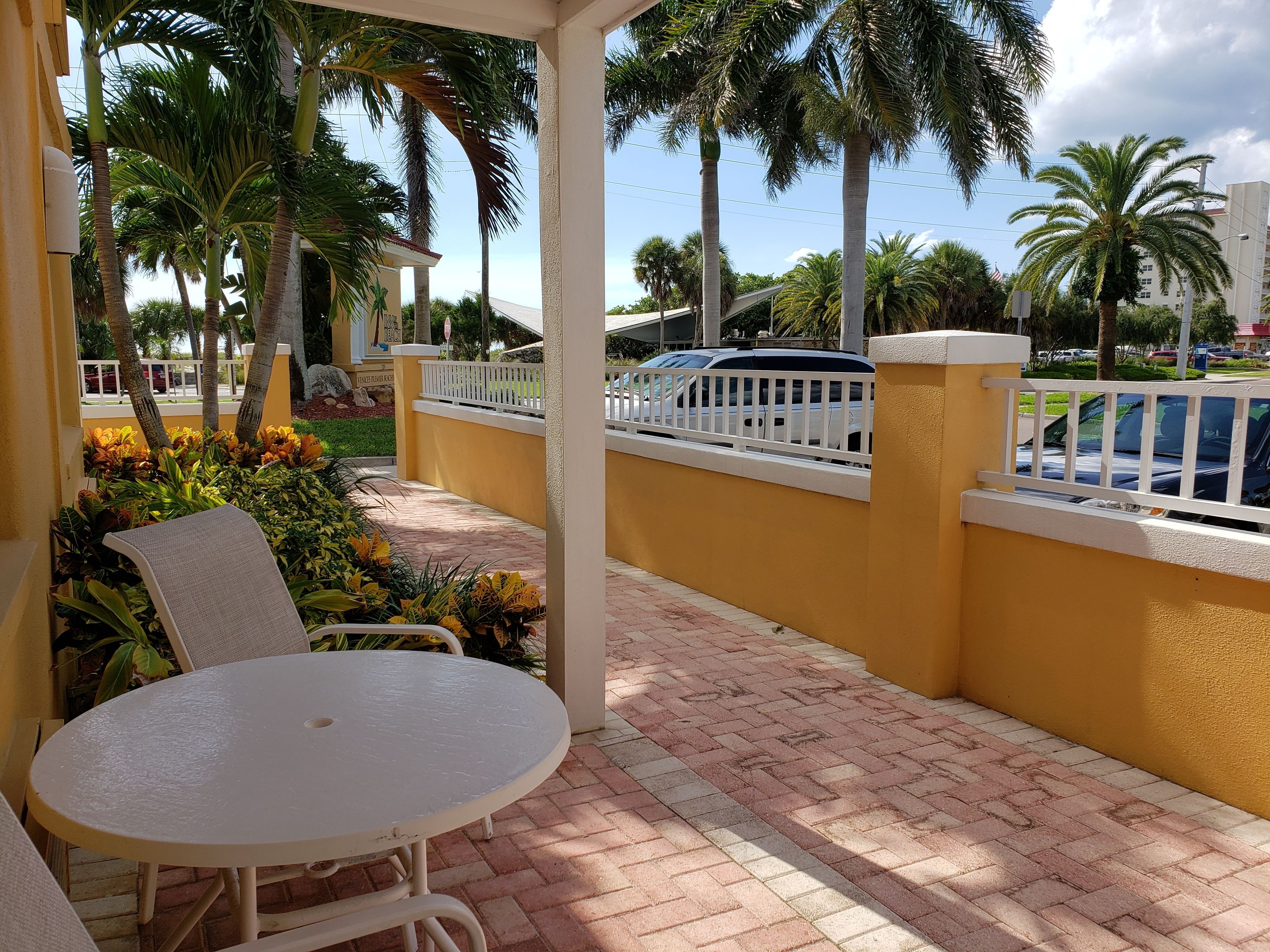 A covered outdoor seating area with a table and chairs, surrounded by a yellow wall and palm trees.