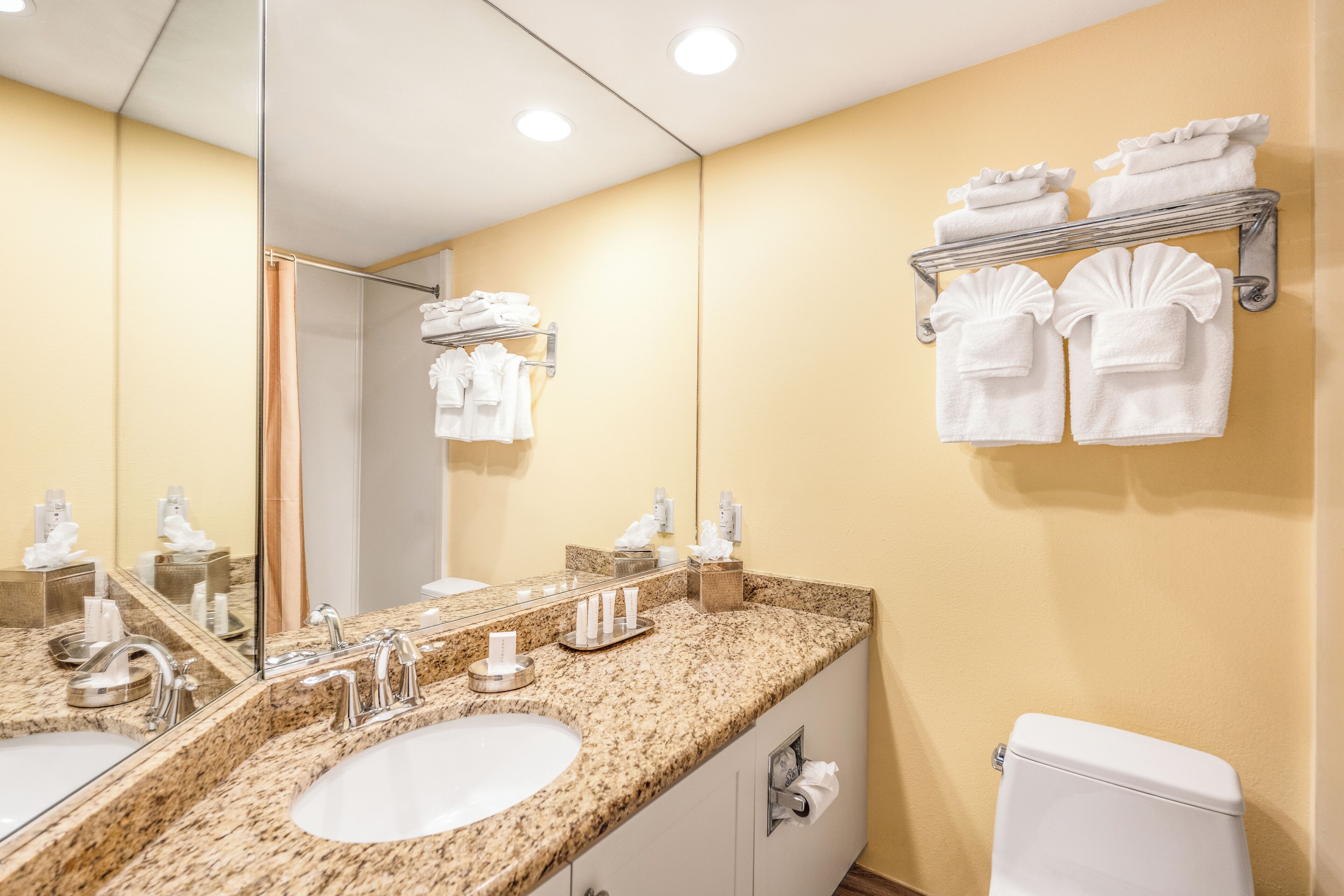 A bathroom with a granite countertop, two sinks, a large mirror, a towel rack, and a toilet.