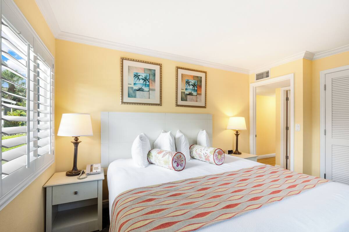 A hotel room with a bed, two nightstands, a window with blinds, two wall-mounted lamps, and two framed pictures.