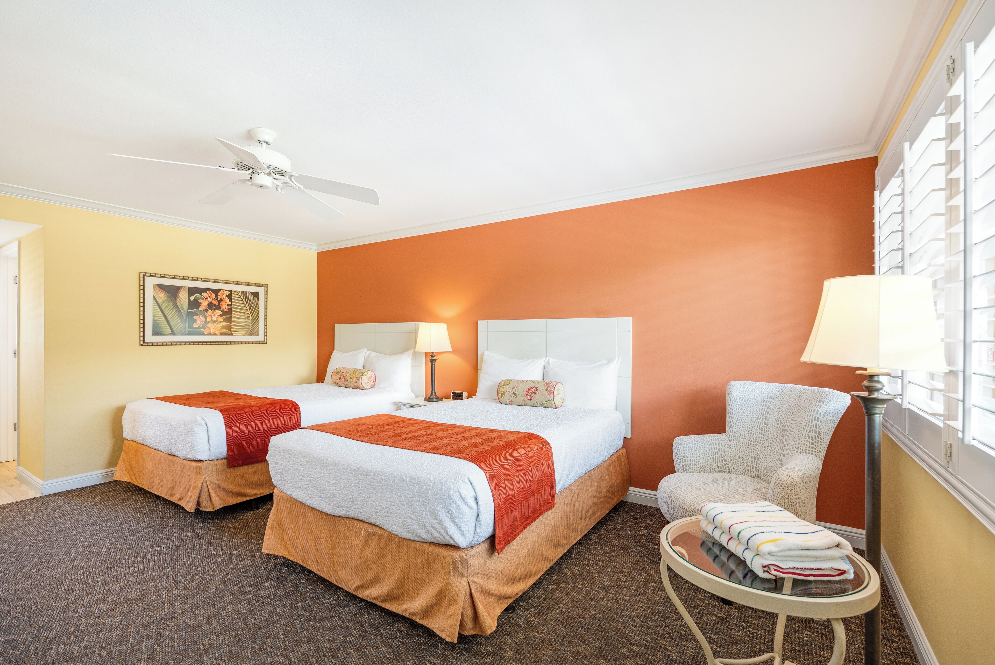 A hotel room with two beds, a ceiling fan, a painting on the wall, and a chair with a table.