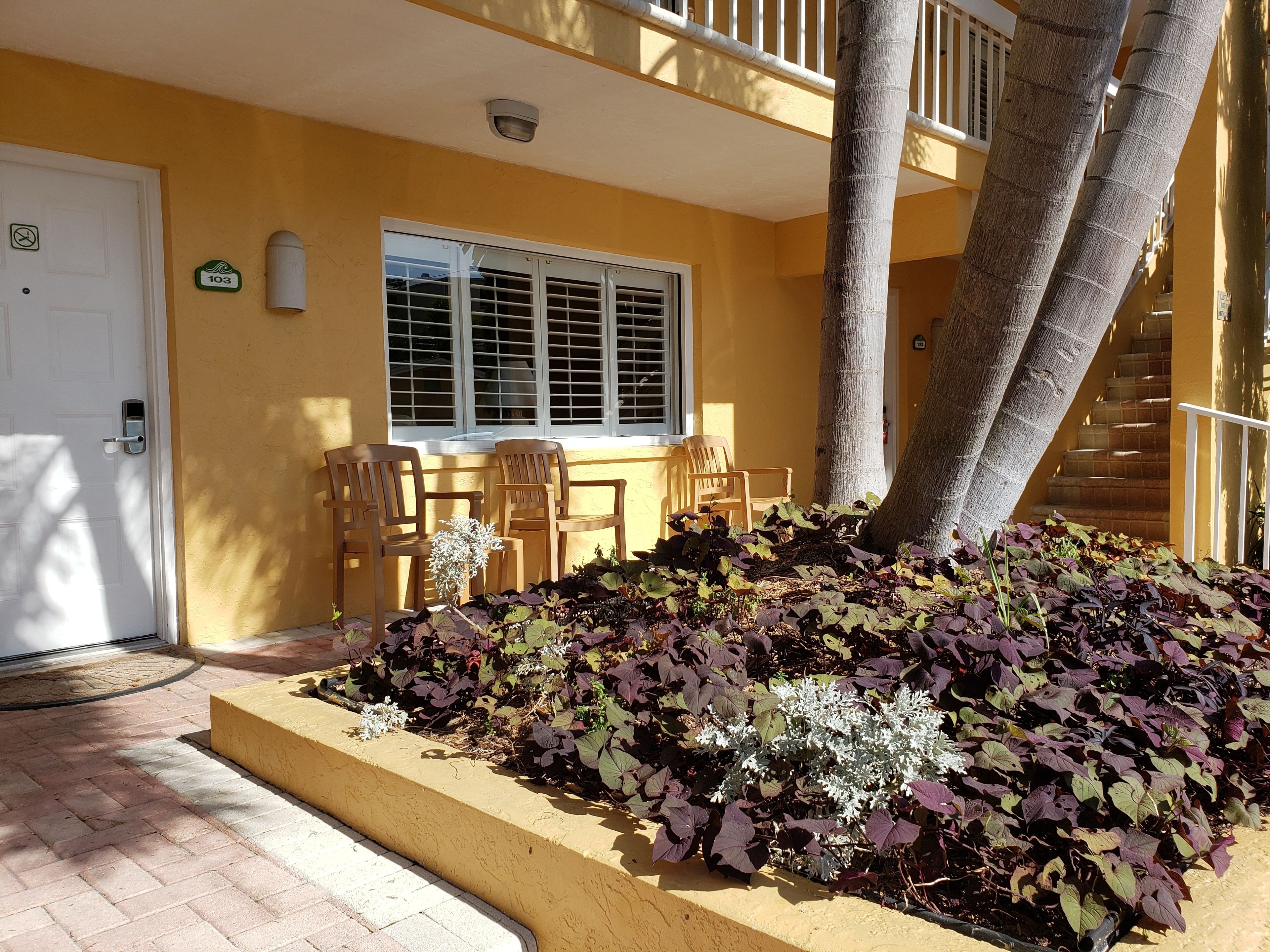 A yellow building with a white door, a window with shutters, and a staircase. There are wooden chairs and a small table with a vase of flowers.