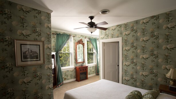 A bedroom with floral wallpaper, a bed with green pillows, a ceiling fan, and a framed picture on the wall. - Maplewood Hotel (Saugatuck)