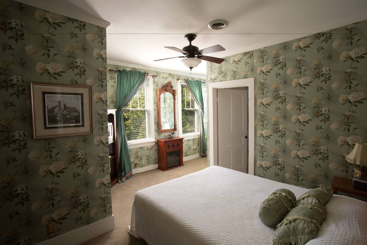 A bedroom with floral wallpaper, a bed with green pillows, a ceiling fan, and a framed picture on the wall.