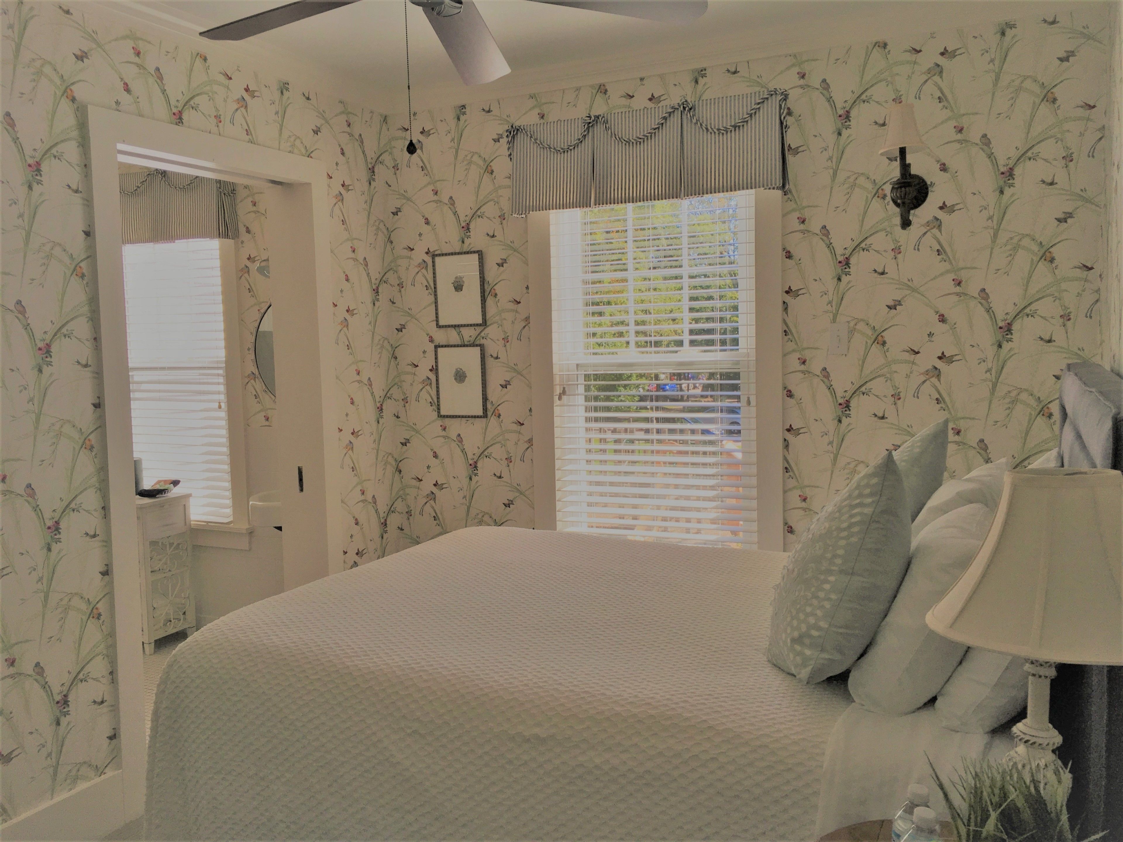 A bedroom with floral wallpaper, a bed with pillows, a bedside table with a lamp, and a window with blinds.