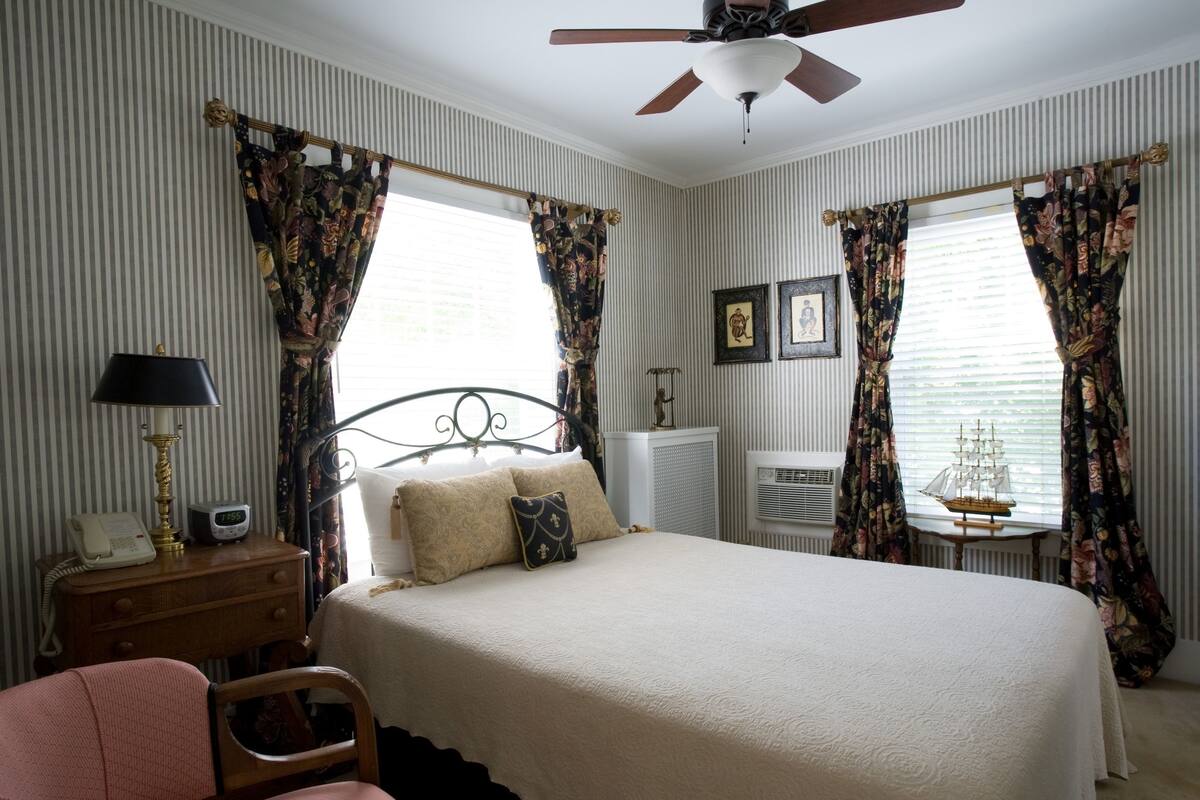 A neatly arranged bedroom with a bed, a chair, a table with a ship model, and curtains.