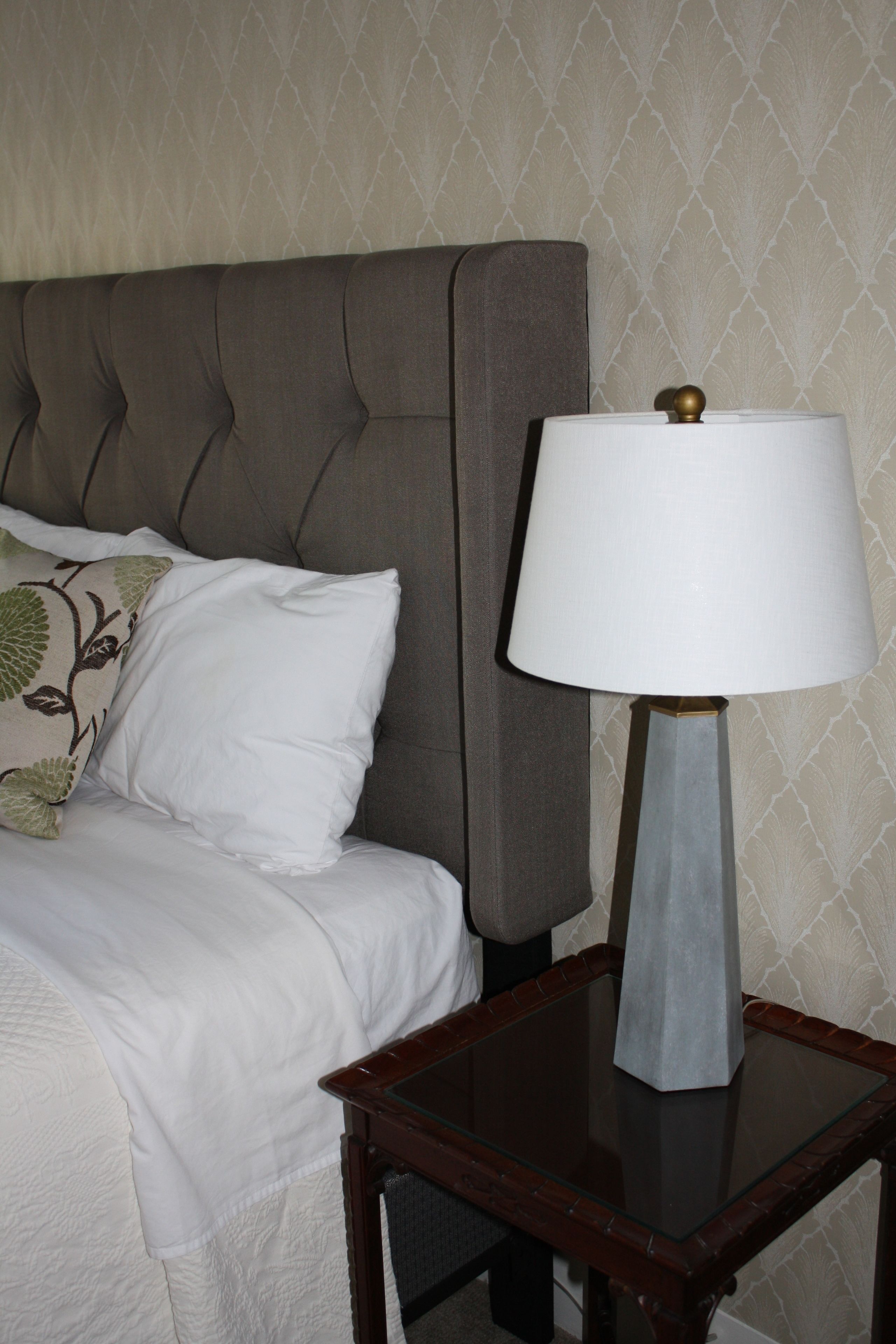 A neatly made bed with a grey headboard, a white pillow, and a bedside table with a lamp.