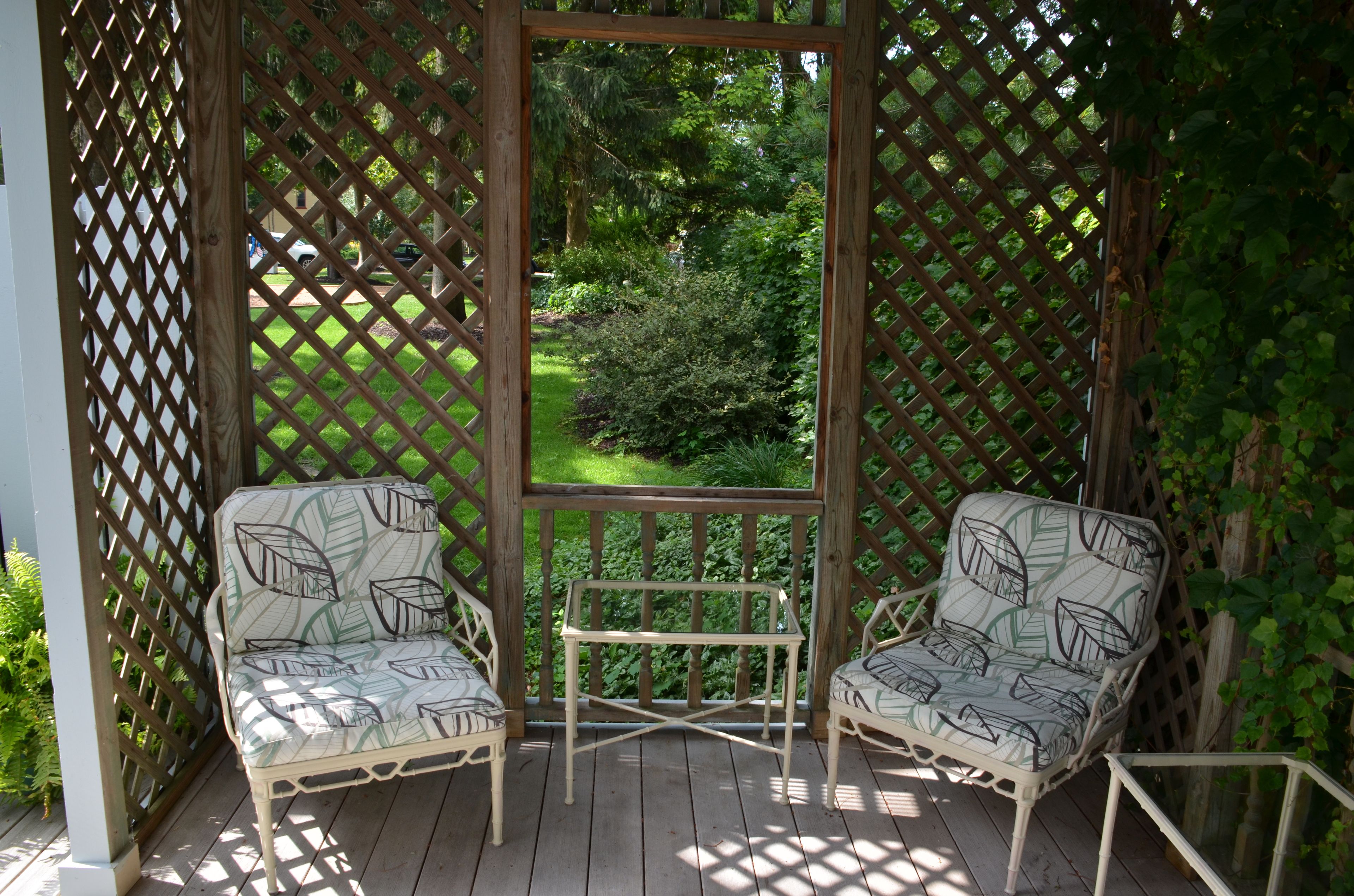 A wooden deck with two patterned chairs and a small table, surrounded by a lattice fence with a view of a garden.