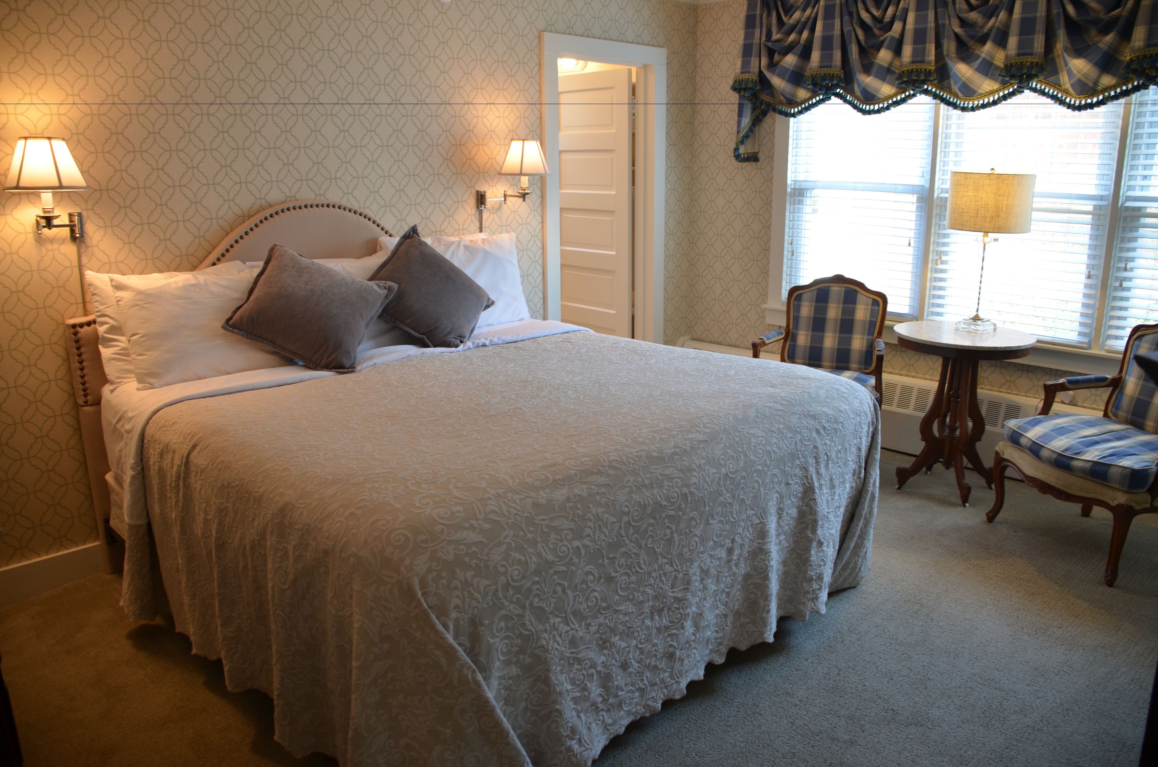 A neatly made bed with a lace bedspread, two bedside lamps, and a plaid chair by a window with a view.