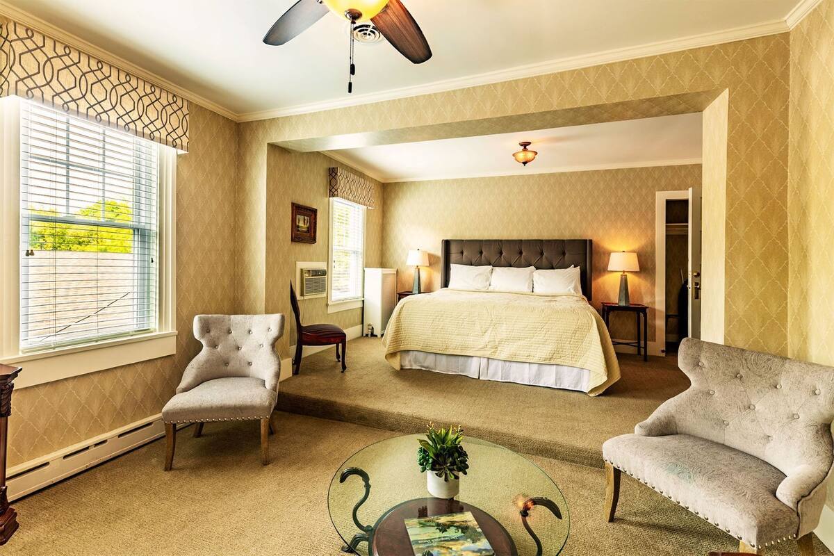 A bedroom with a large bed, two armchairs, a glass coffee table, and a ceiling fan.