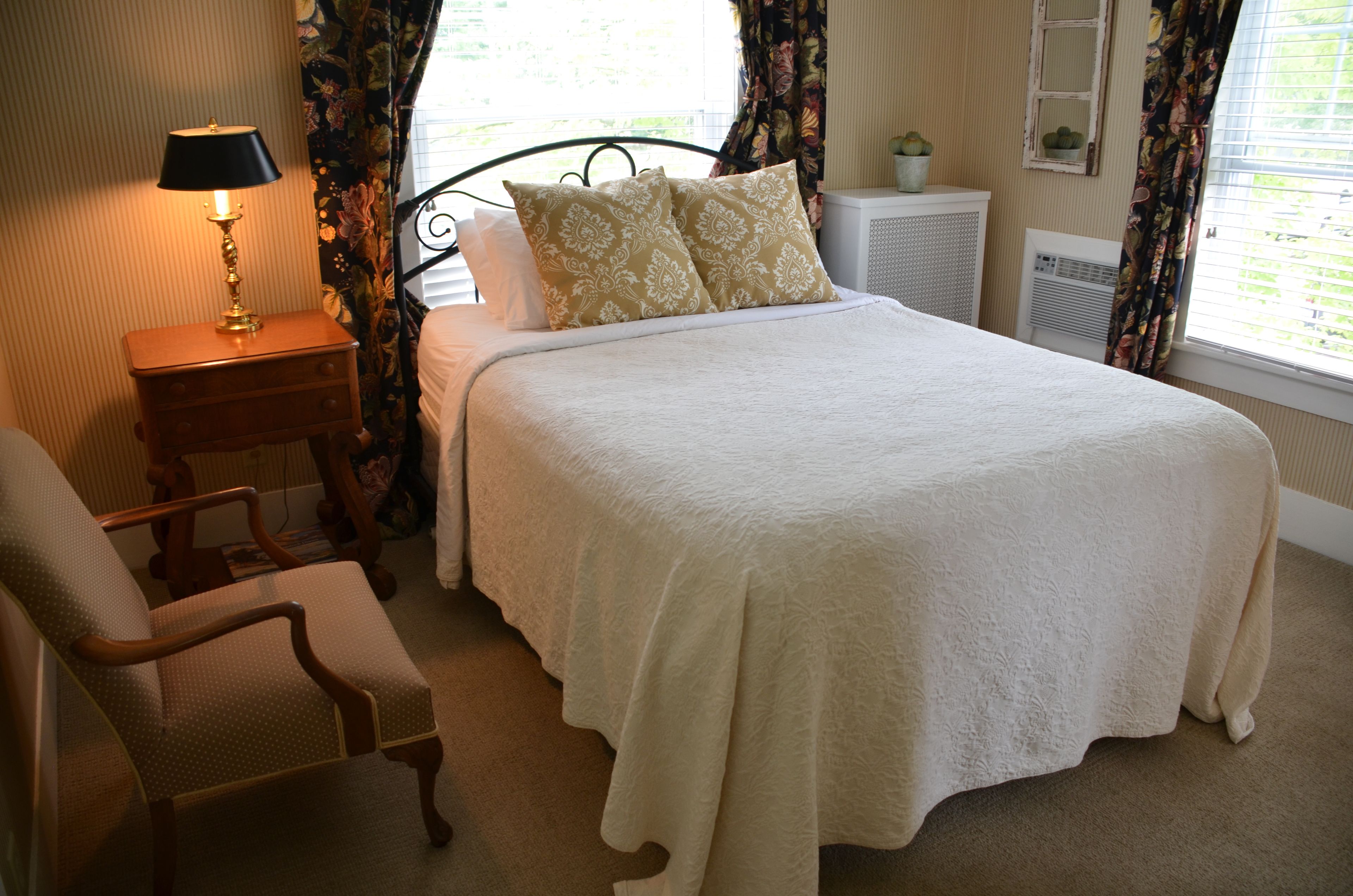 A neatly made bed with a white bedspread and two decorative pillows, a wooden nightstand with a lamp, a chair, and a radiator.