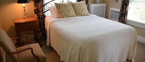 A neatly made bed with a white bedspread and two decorative pillows, a wooden nightstand with a lamp, a chair, and a radiator.