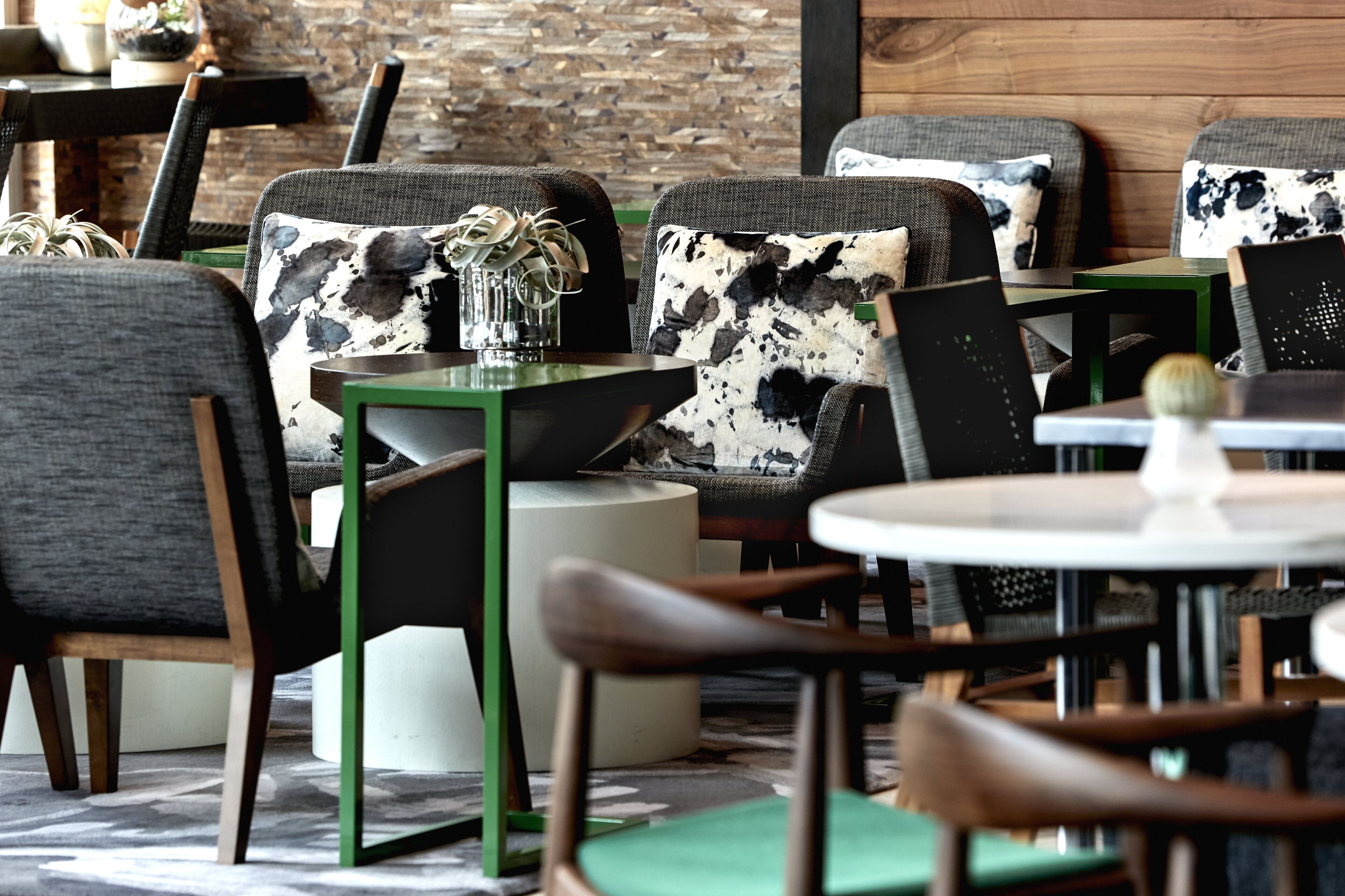 A modern restaurant interior with a mix of grey and black upholstered chairs, a green table, and a rustic brick wall in the background.