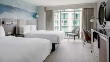 A hotel room with two beds, a desk, a chair, a TV, and a large window with a view of buildings.