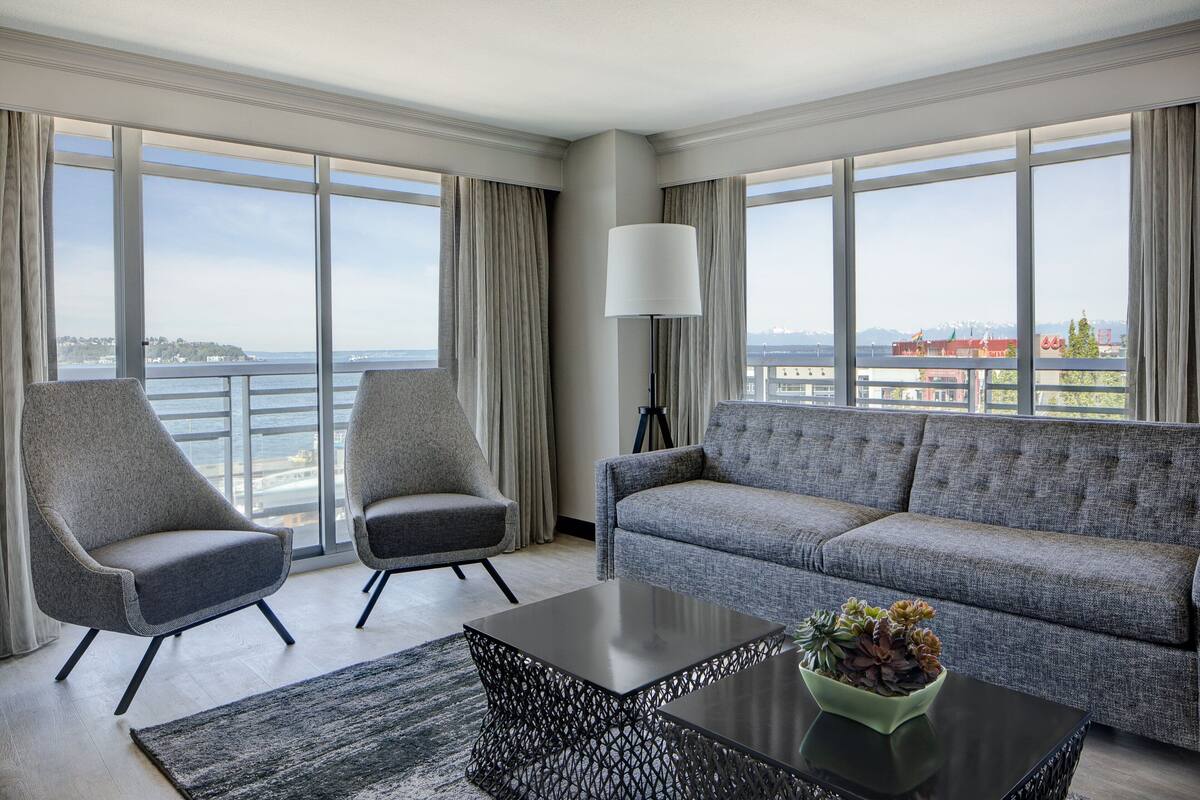 A modern hotel room with a large window, a grey sofa, two armchairs, a coffee table, and a floor lamp.