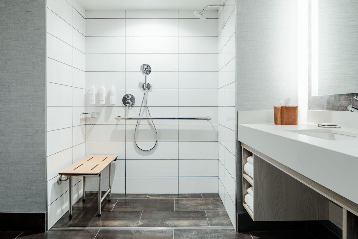 A modern bathroom with white tiled walls, a shower with a handheld showerhead, a wooden bench, and a white sink with a towel rack.