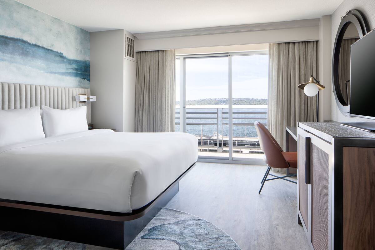 A hotel room with a large bed, a desk with a TV, a view of the sea through the window, and a balcony.