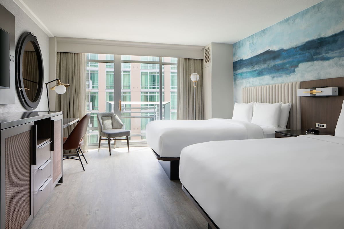 A hotel room with two beds, a desk with a chair, a large window with a view of buildings, and a painting on the wall.