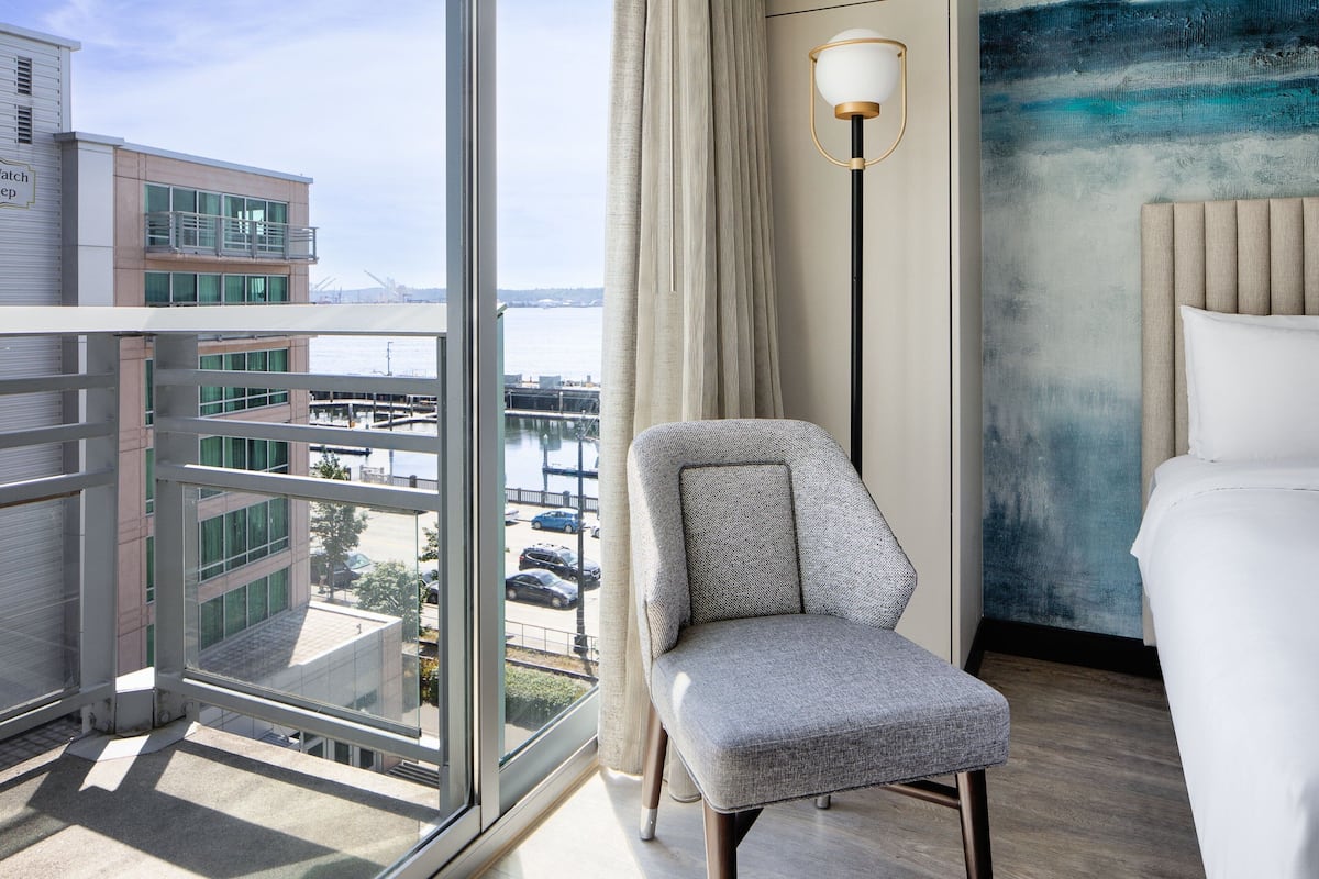 A hotel room with a large window, a chair, a bed, and a painting on the wall.