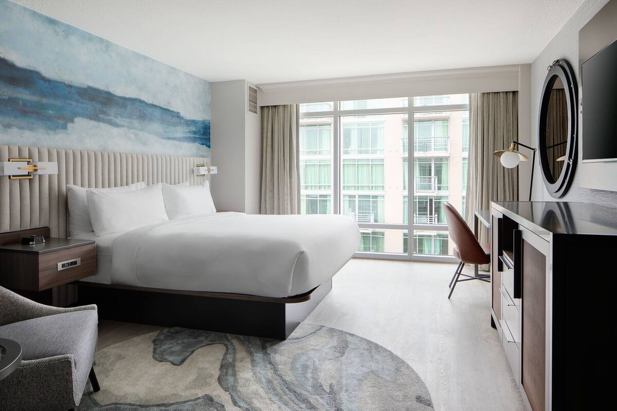 A modern hotel room with a large bed, a desk, a chair, and a view of the city through large windows.