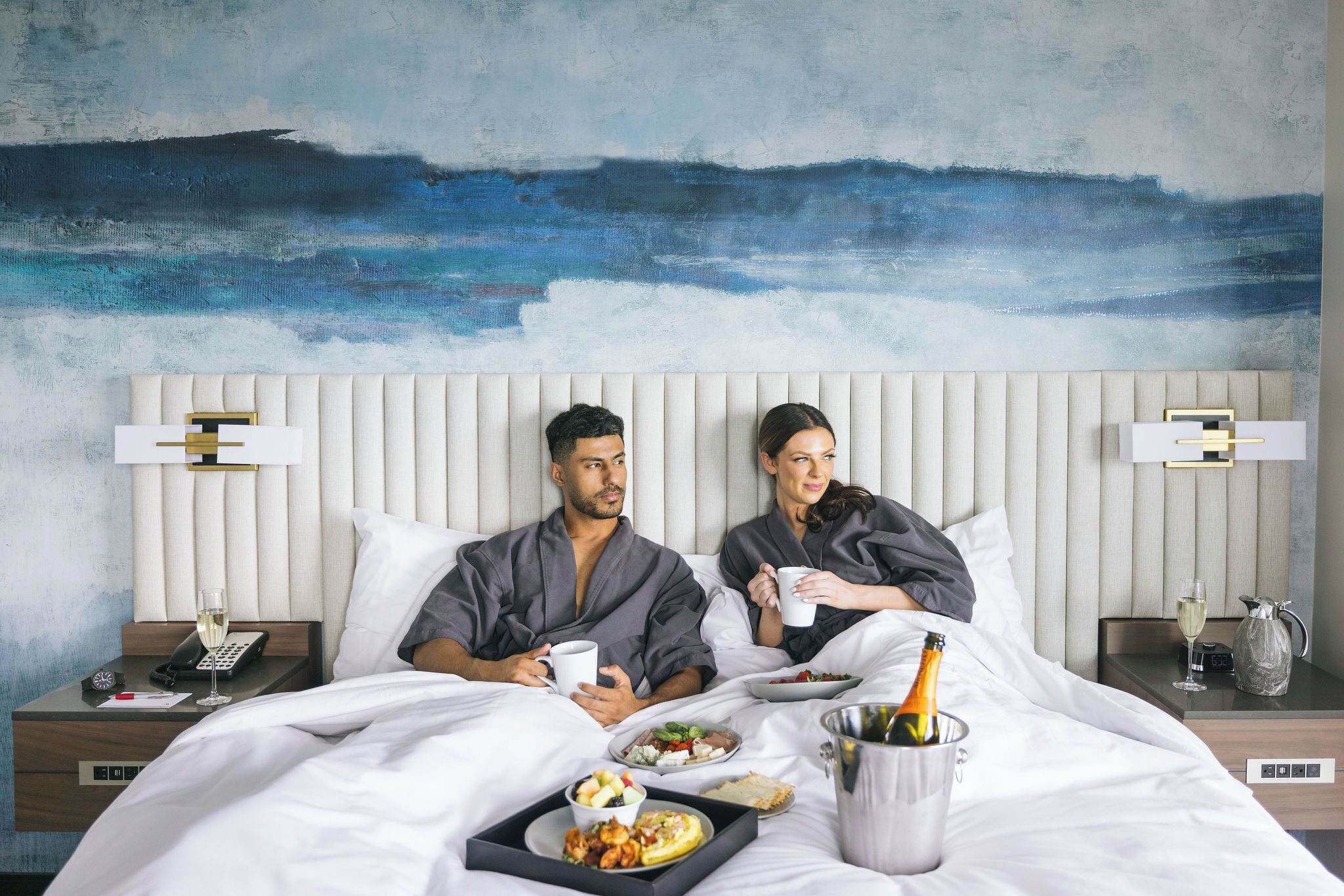A hotel room with a bed, bedside tables, a phone, a bottle of champagne, and a painting on the wall.