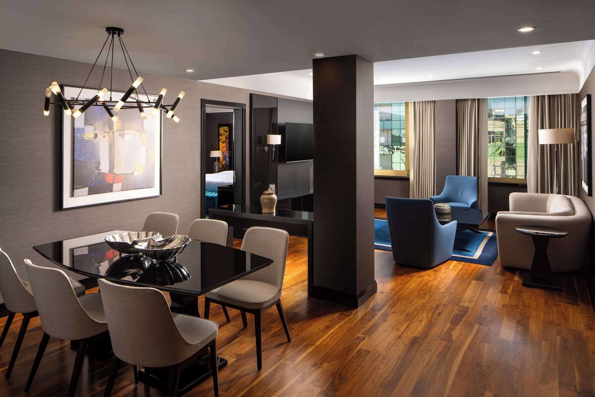 A modern hotel room with a dining area, a living space with a sofa and armchairs, a TV, and a large window offering a city view.