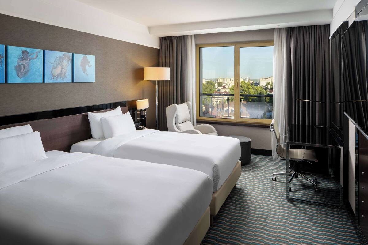 A hotel room with two beds, a desk, a chair, a lamp, and a large window with a city view.