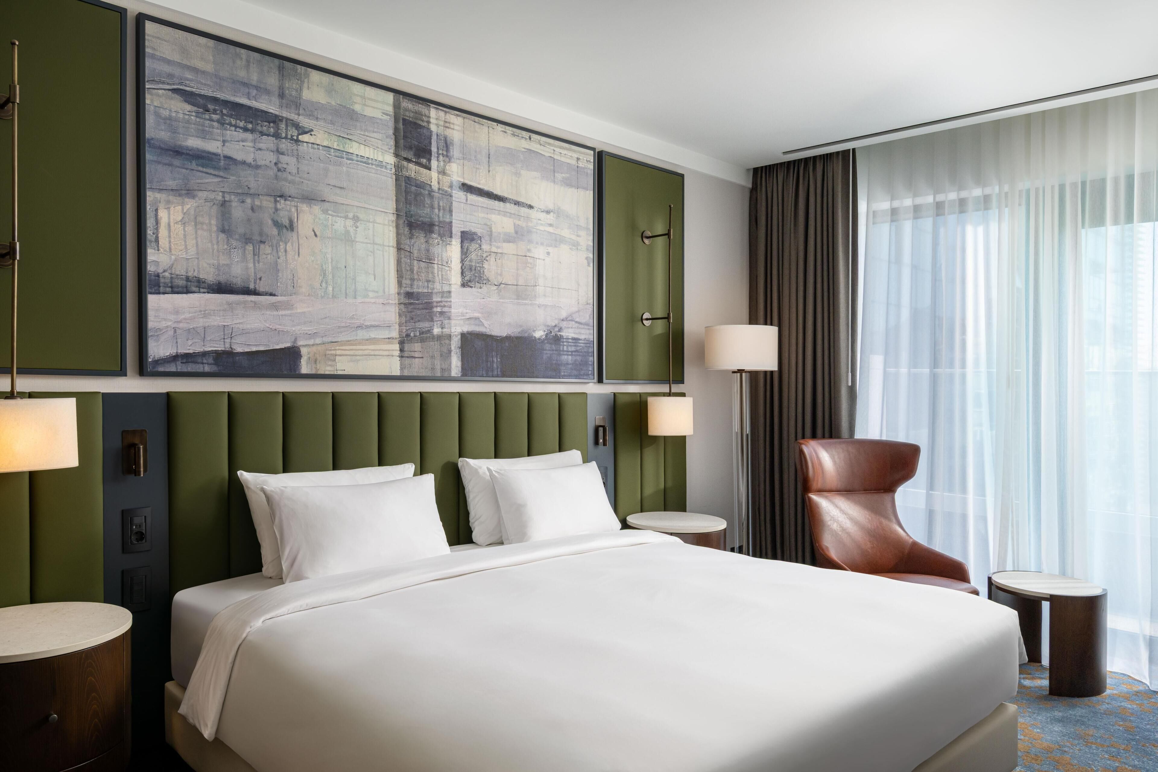 A hotel room with a large bed, a headboard with vertical panels, a wall-mounted artwork, a round side table, a chair, and a large window with curtains.