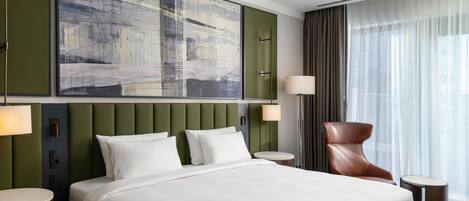 A hotel room with a large bed, a headboard with vertical panels, a wall-mounted artwork, a round side table, a chair, and a large window with curtains.