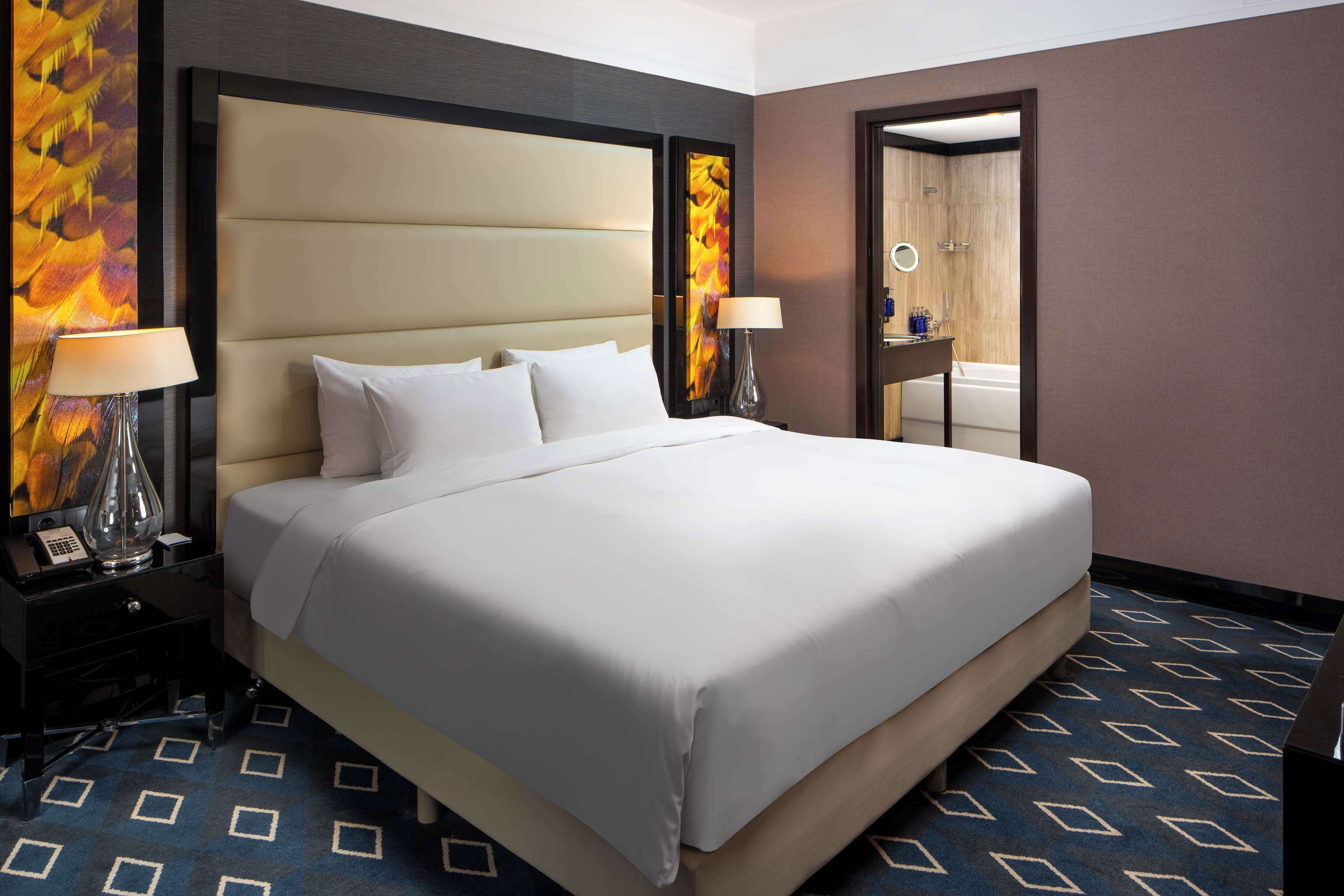 A hotel room with a large bed, two bedside tables with lamps, a mirror, and a bathroom visible through an open door.