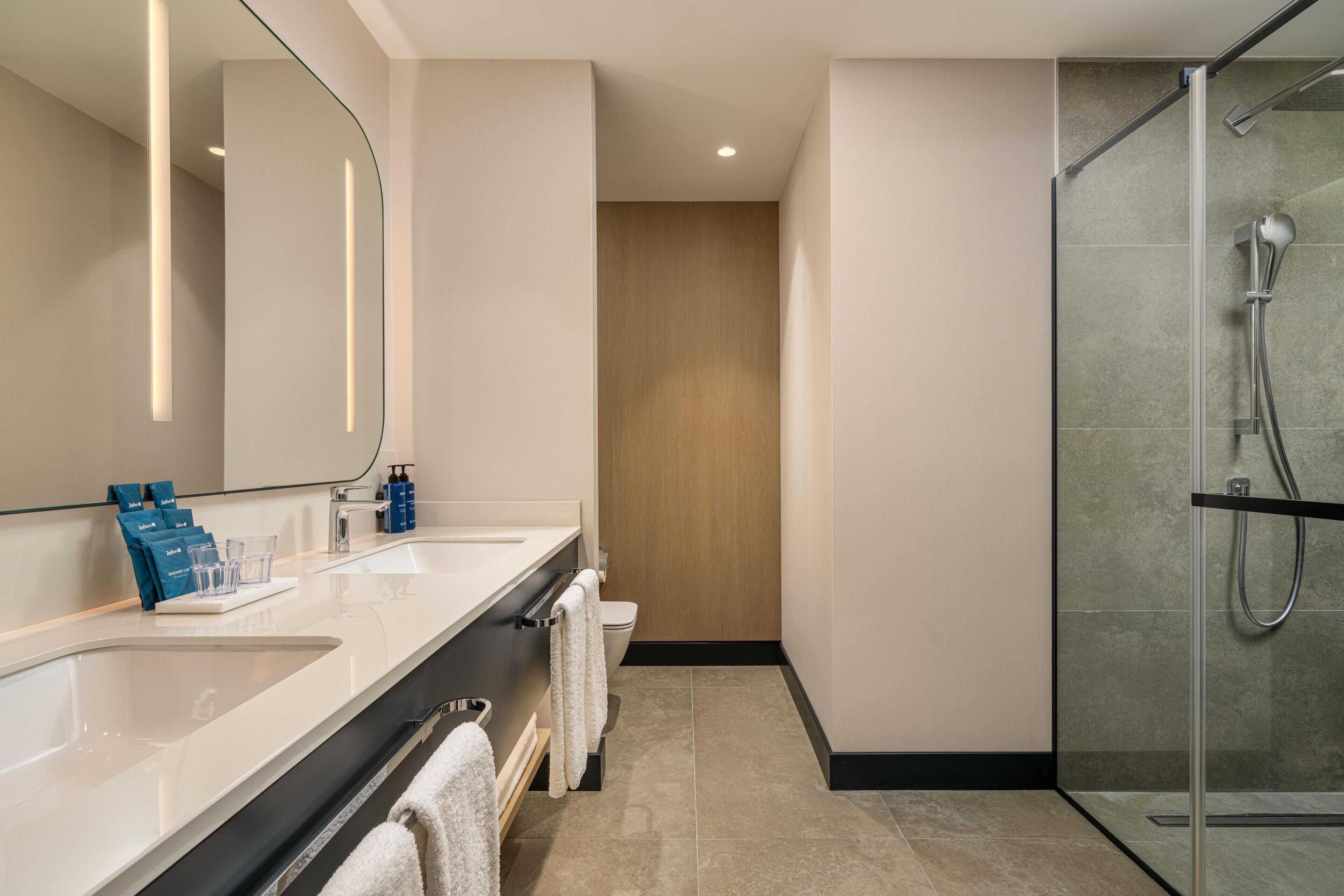 A modern bathroom with two sinks, a glass shower enclosure, and a wooden door in the background.
