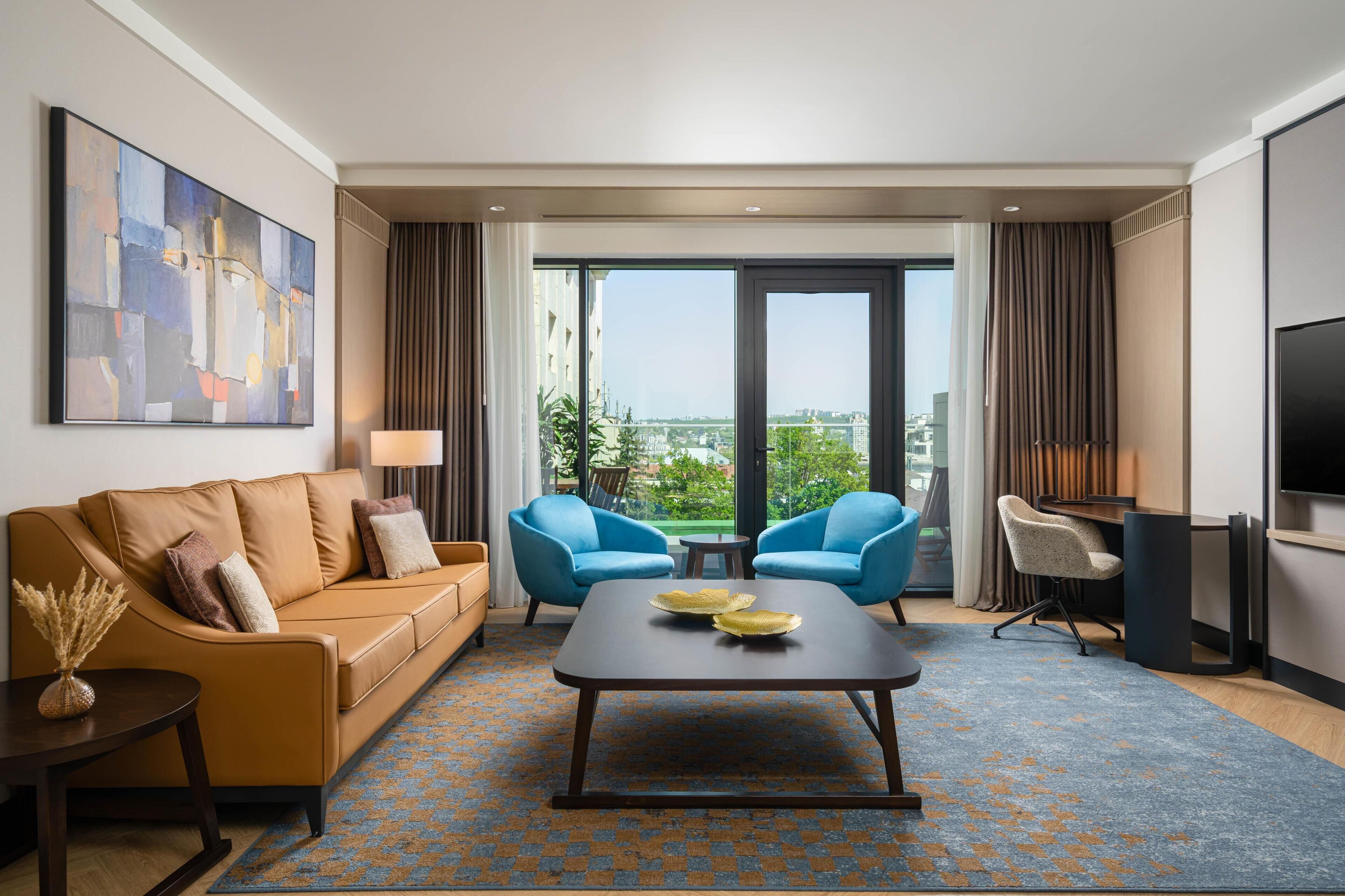 Une chambre d’hôtel moderne équipée d’un canapé, de deux fauteuils, d’une table basse, d’un téléviseur et d’une grande fenêtre donnant sur la ville.
