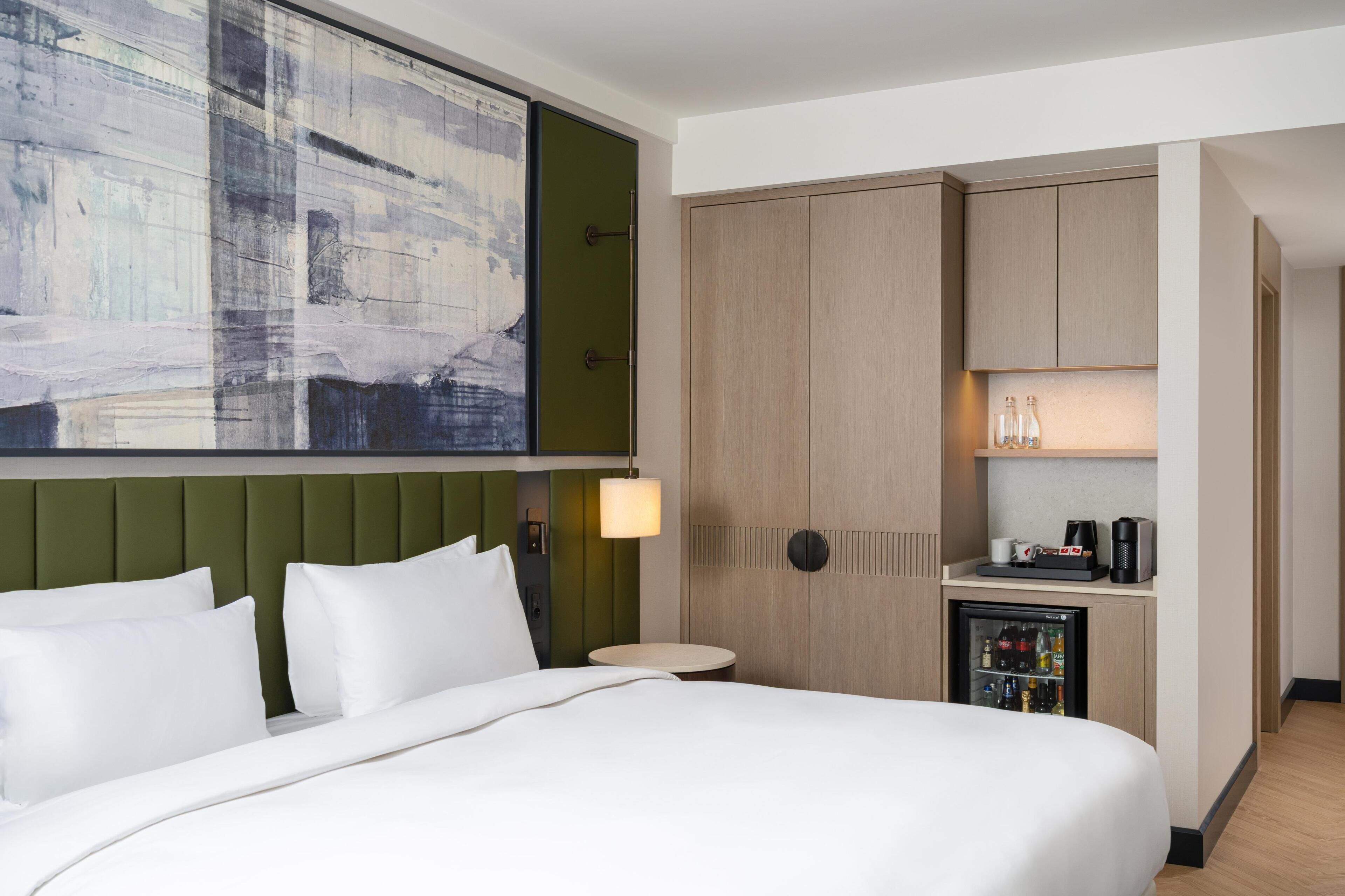 A hotel room with a large bed, a green headboard, a wall-mounted painting, a minibar, and a small table with a lamp.