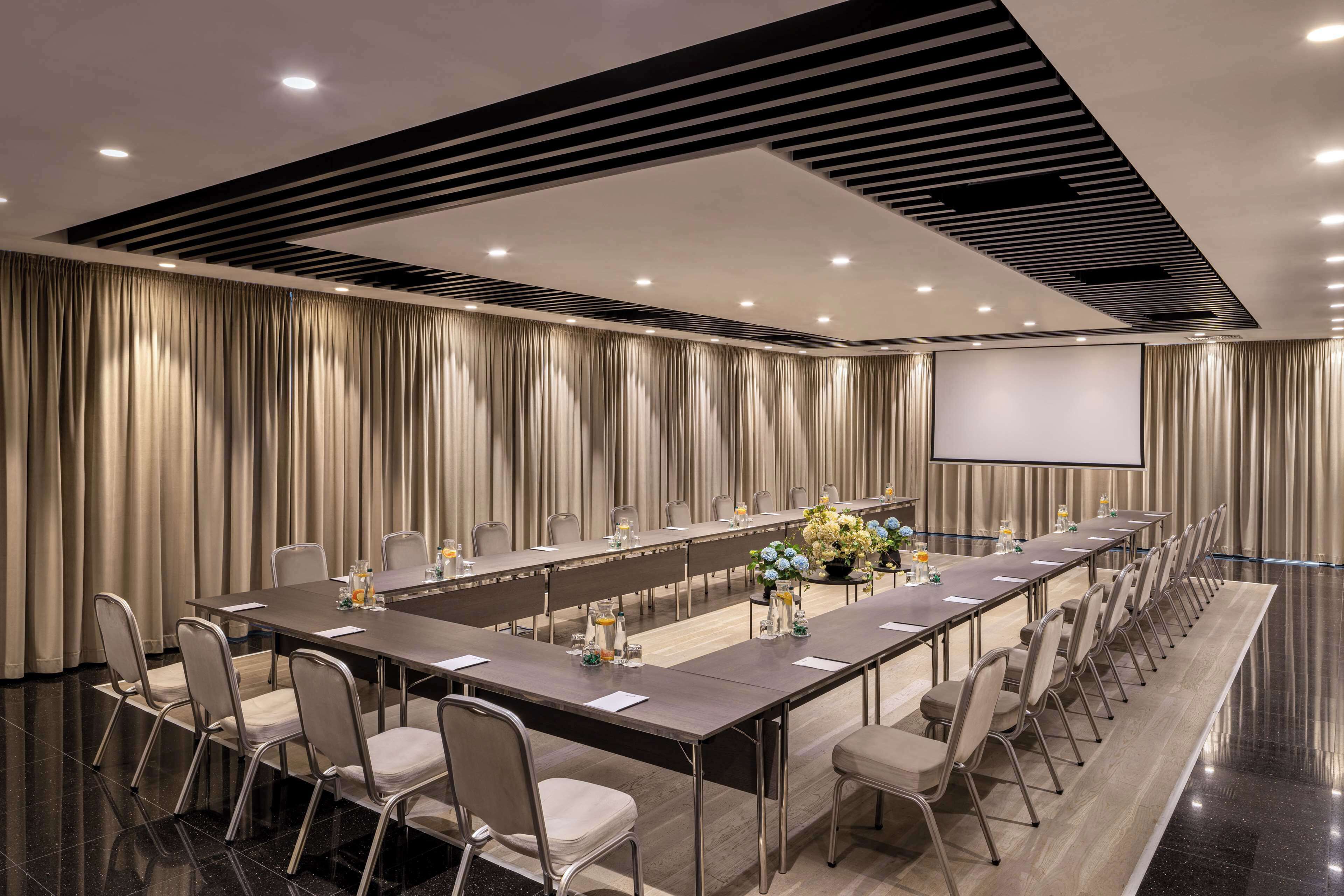 Un salón de conferencias con una larga mesa, sillas, una pantalla de proyector y un arreglo floral.