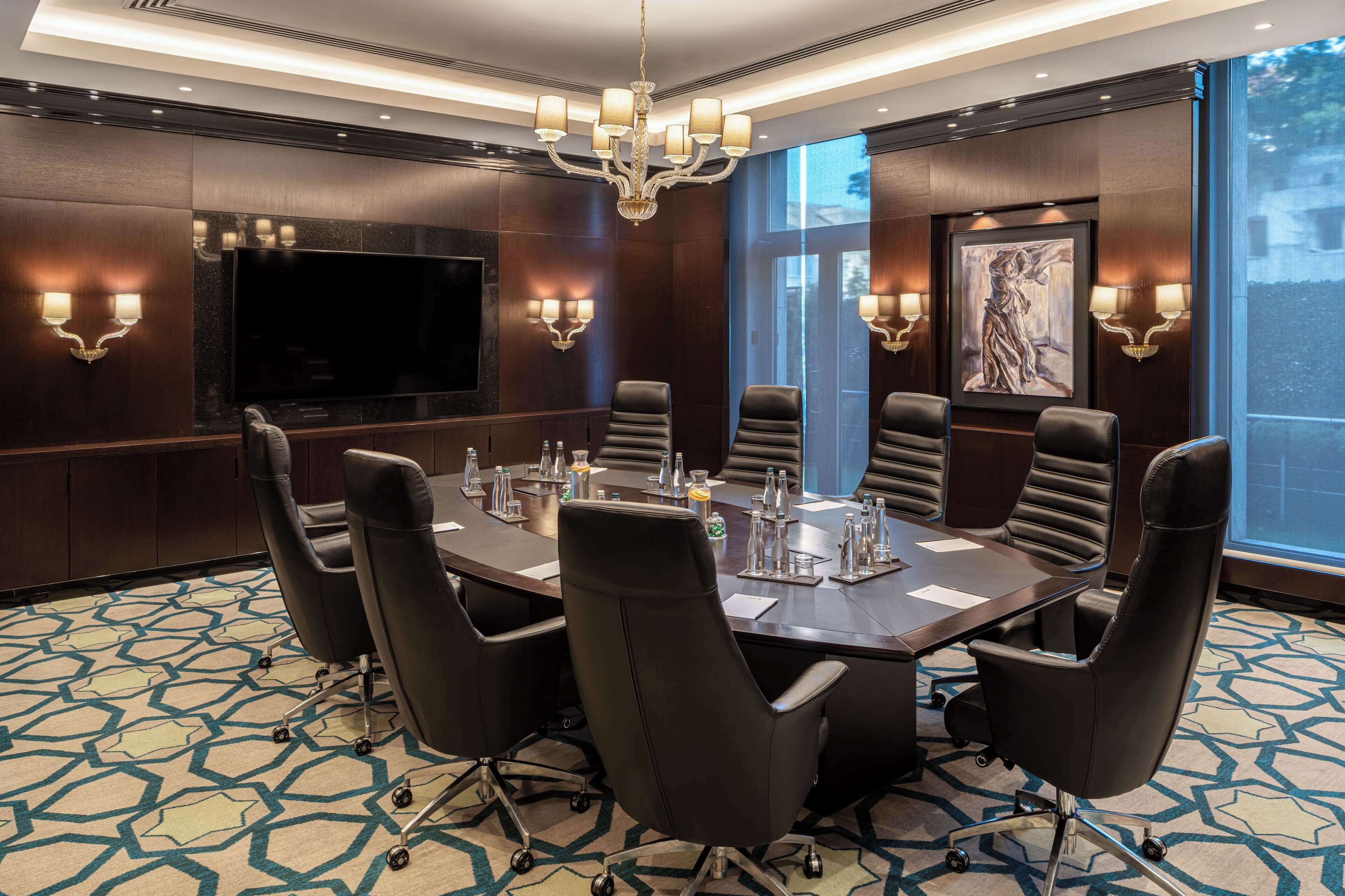 A conference room with a large table, black leather chairs, a chandelier, wall-mounted TV, and artwork.
