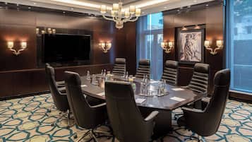 A conference room with a large table, black leather chairs, a chandelier, wall-mounted TV, and artwork.