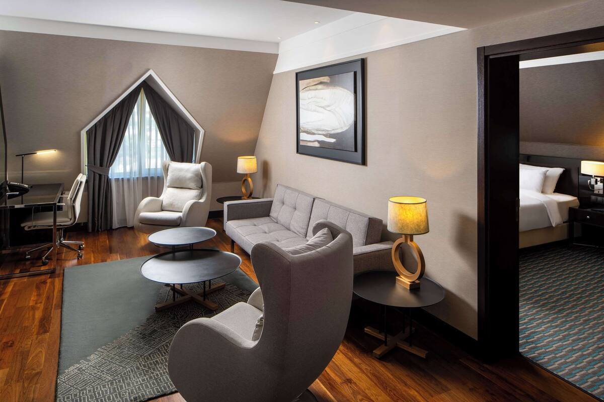 A modern hotel room with a sofa, armchair, and a small round table. There is a bed with a headboard and a nightstand with a lamp.