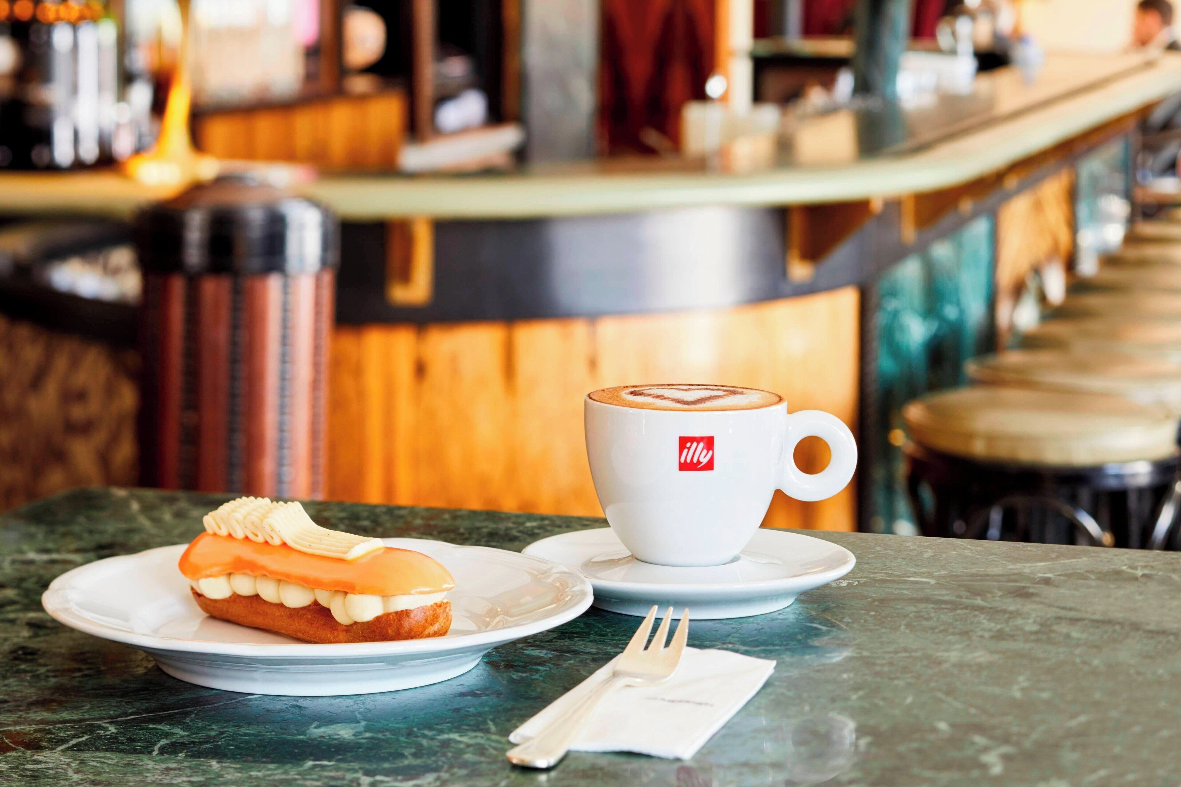 Een tafel met een gebakje, een kop koffie, een servetje, en een toonbank op de achtergrond.