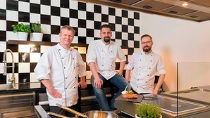 Tres chefs en una cocina moderna con salpicadero de cuadros, electrodomésticos de acero inoxidable y plantas en macetas.