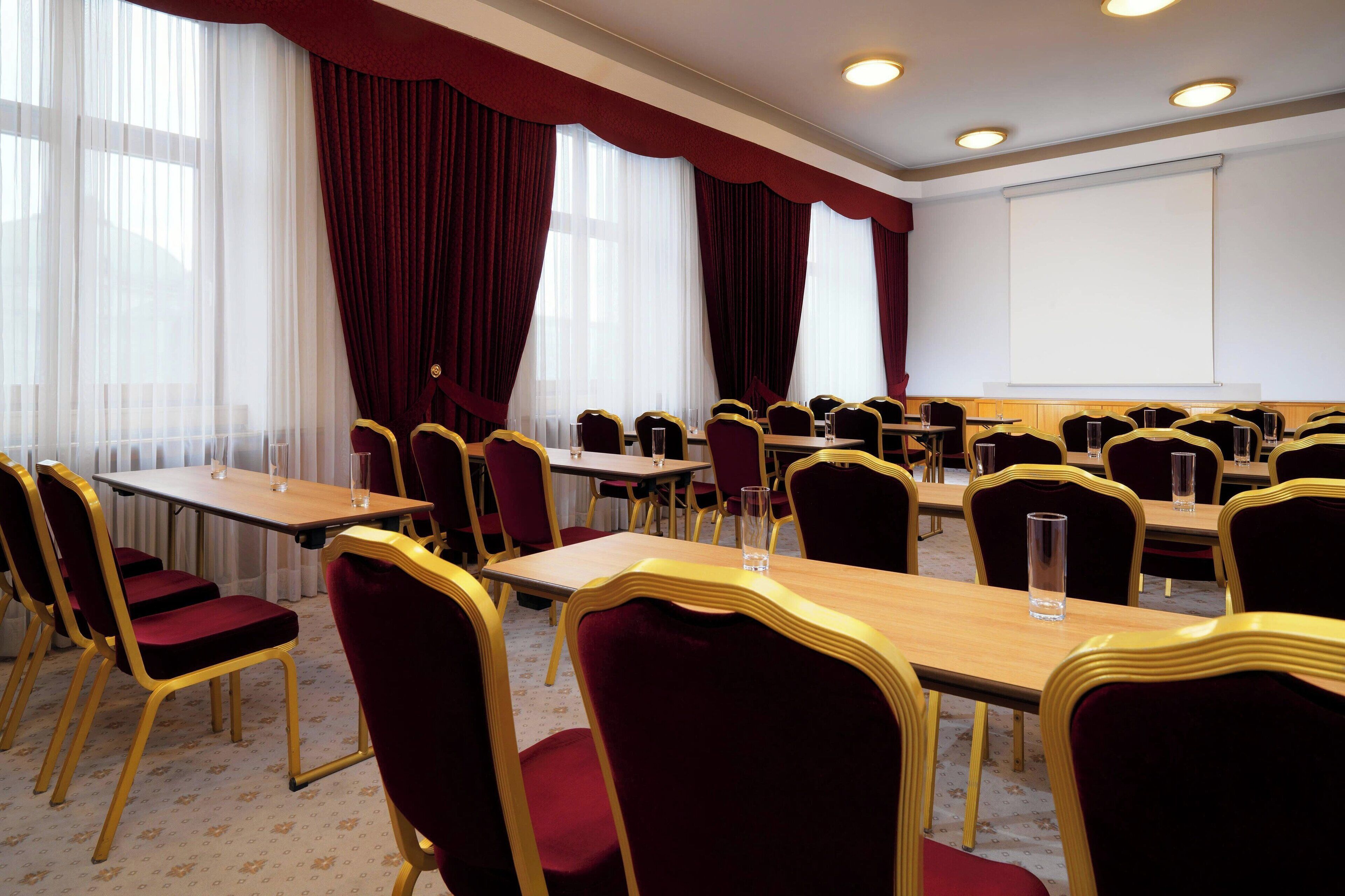 A conference room with multiple tables and chairs, a projector screen, and large windows with curtains.