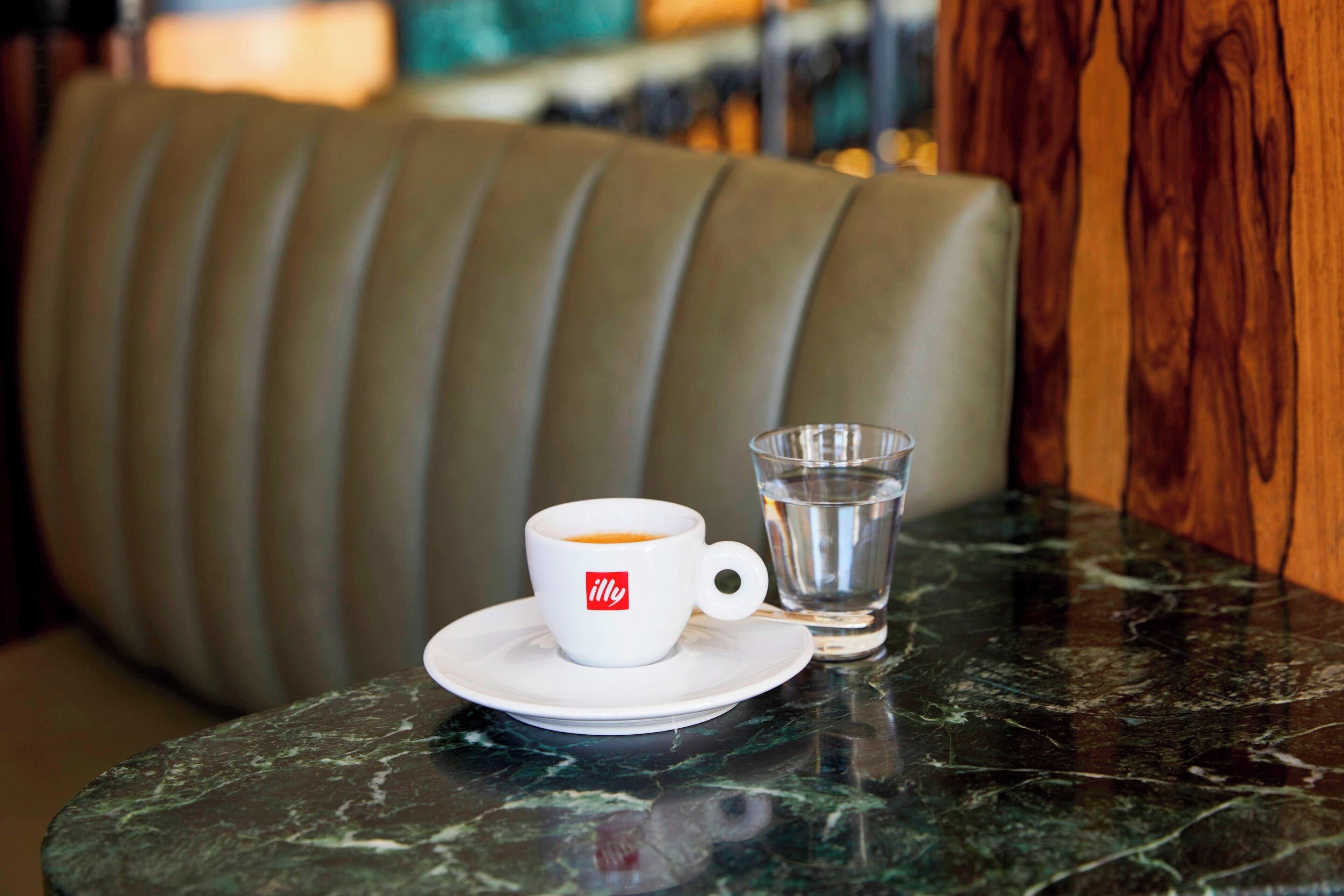 A white cup with a logo on a saucer, a glass of water, and a marble table in a restaurant setting.