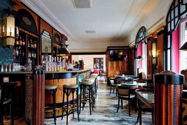 A bar area with a marble floor, wooden bar counter, and stools. There are tables and chairs in the background.