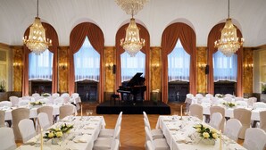Un salón de baile elegante con un escenario, candelabros y un piano de cola. Mesas dispuestas con manteles blancos y sillas.