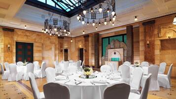 Un salón de banquetes con mesas redondas, manteles blancos y sillas, que cuenta con un techo alto con candelabros y una chimenea de piedra.