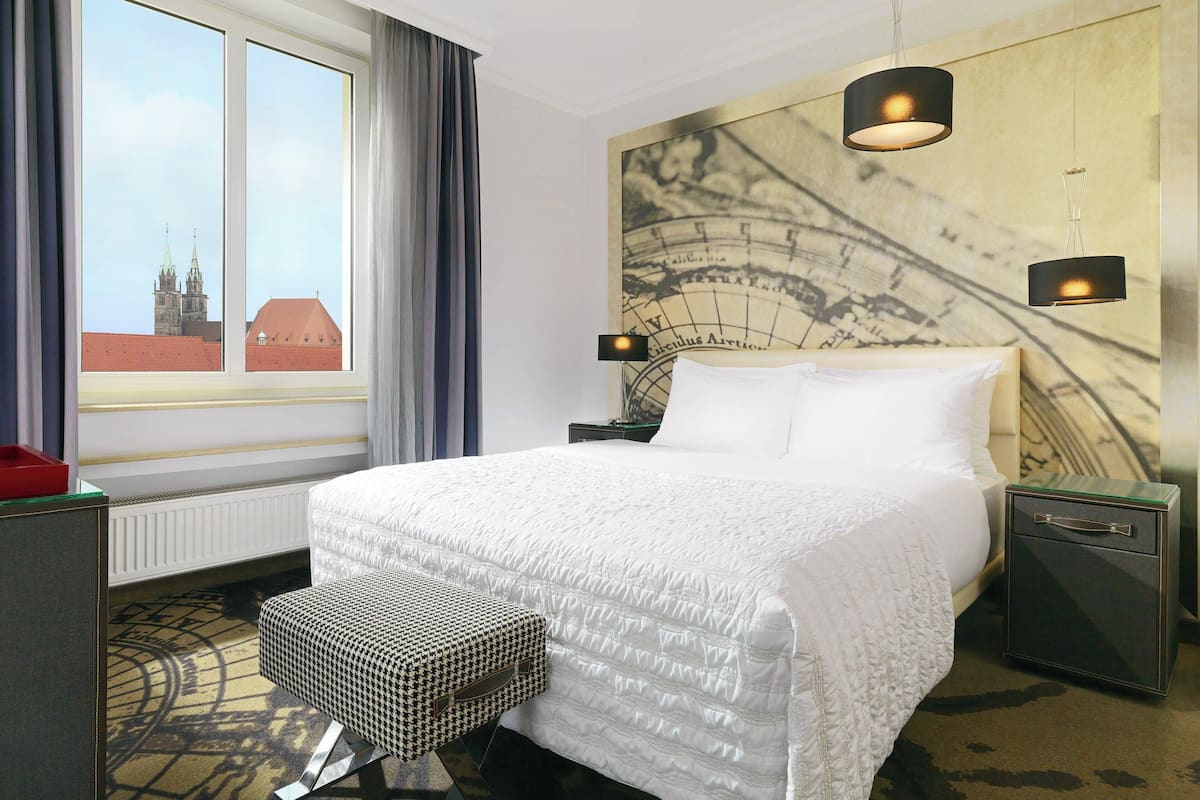 Une chambre d’hôtel avec un grand lit, une table de chevet avec une lampe, une petite table, un banc et une vue sur le paysage urbain par la fenêtre.