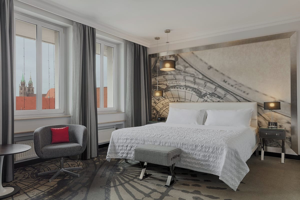 Une chambre d’hôtel avec un grand lit, une chaise, une petite table et une vue sur le paysage urbain par les fenêtres.